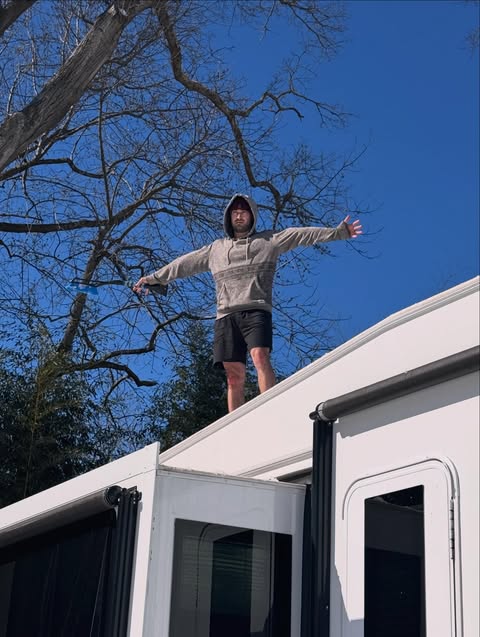 Zach in a beige hoodie and black shorts stands with arms outstretched on top of a white recreational vehicle against a bright blue sky and bare tree branches.