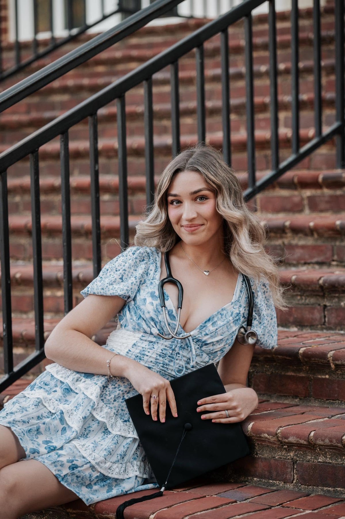A woman with blonde wavy hair sits on brick stairs behind a black railing, wearing a light blue and white floral print dress with a stethoscope around her neck and holding a black clutch bag.