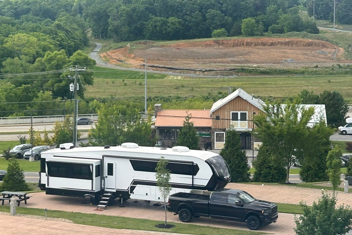 A white motorhome with a black pickup truck parked at a campground, with a small cabin, trees, and an open hillside in the background.