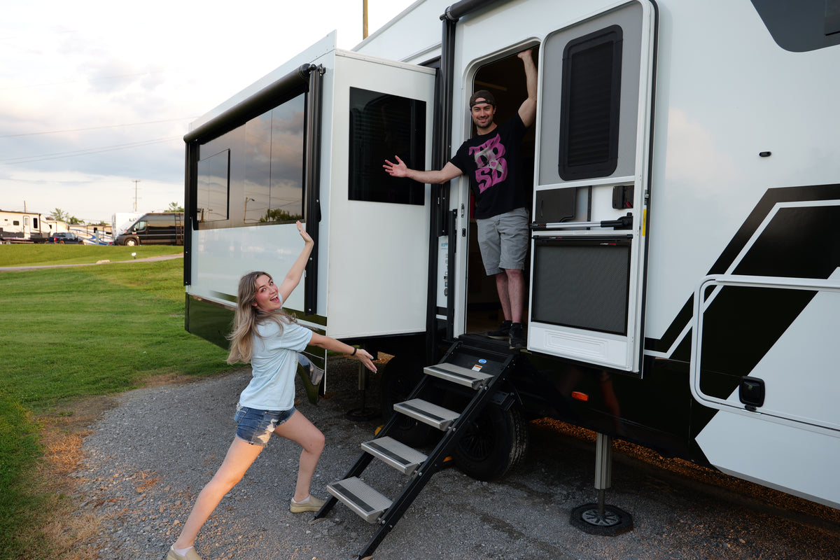 Zach and Ryan pose with a white and black travel trailer at a campground. Zach stands in the open doorway of the trailer, while Ryan poses outside on the gravel ground next to the trailer steps.