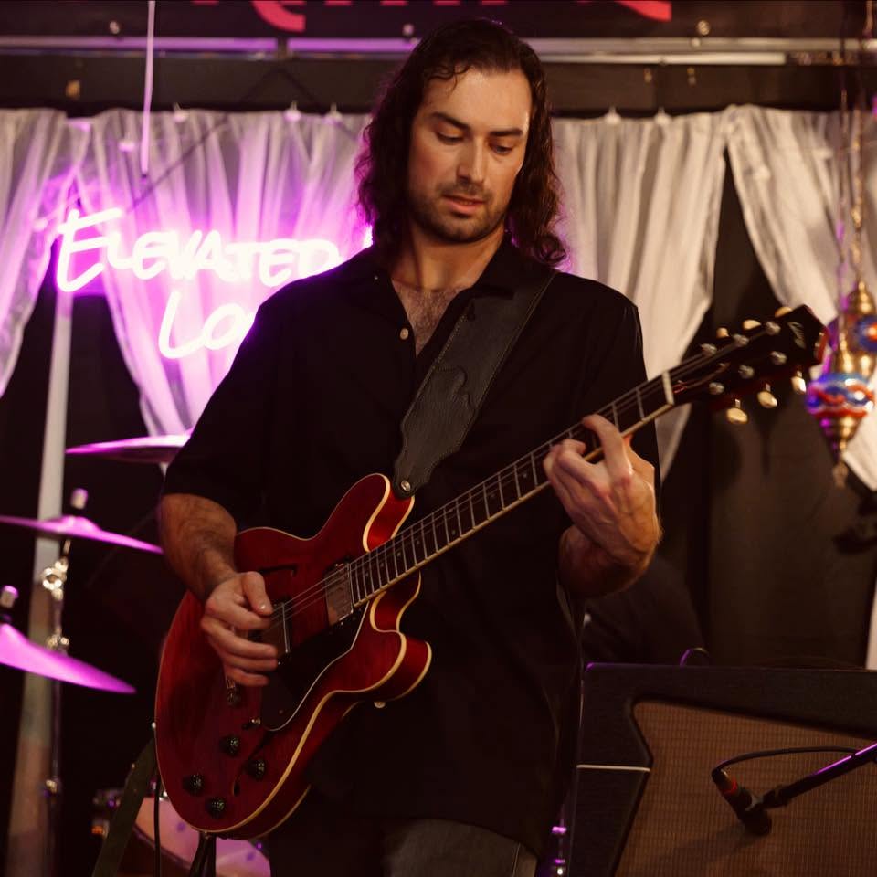 A man with long dark hair playing a red semi-hollow electric guitar on a stage, wearing a black shirt. A pink neon sign and white draped curtains are visible in the background.