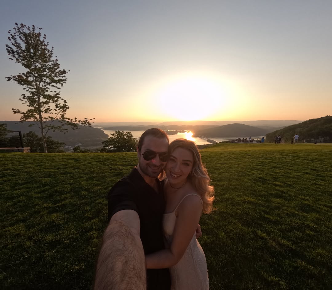 A couple taking a selfie outdoors at sunset, with Ryan in a light dress and Zach in a dark shirt, set against a scenic landscape with hills, water, and the sun low on the horizon.